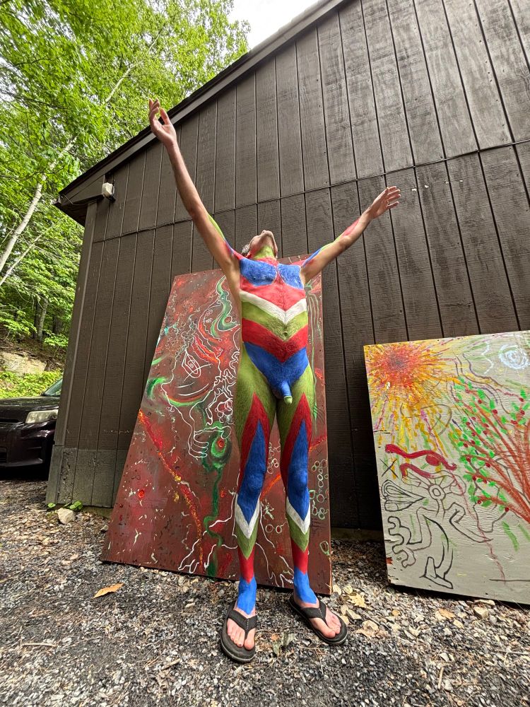 My body painted from my neck to the top of my feet, with blue, red, white, and green patterns. I’m wearing only sandals and my arms and face are reaching for the sky. Behind and next to me are two canvasses with casual painted designs on them. They lean against a brown building.  