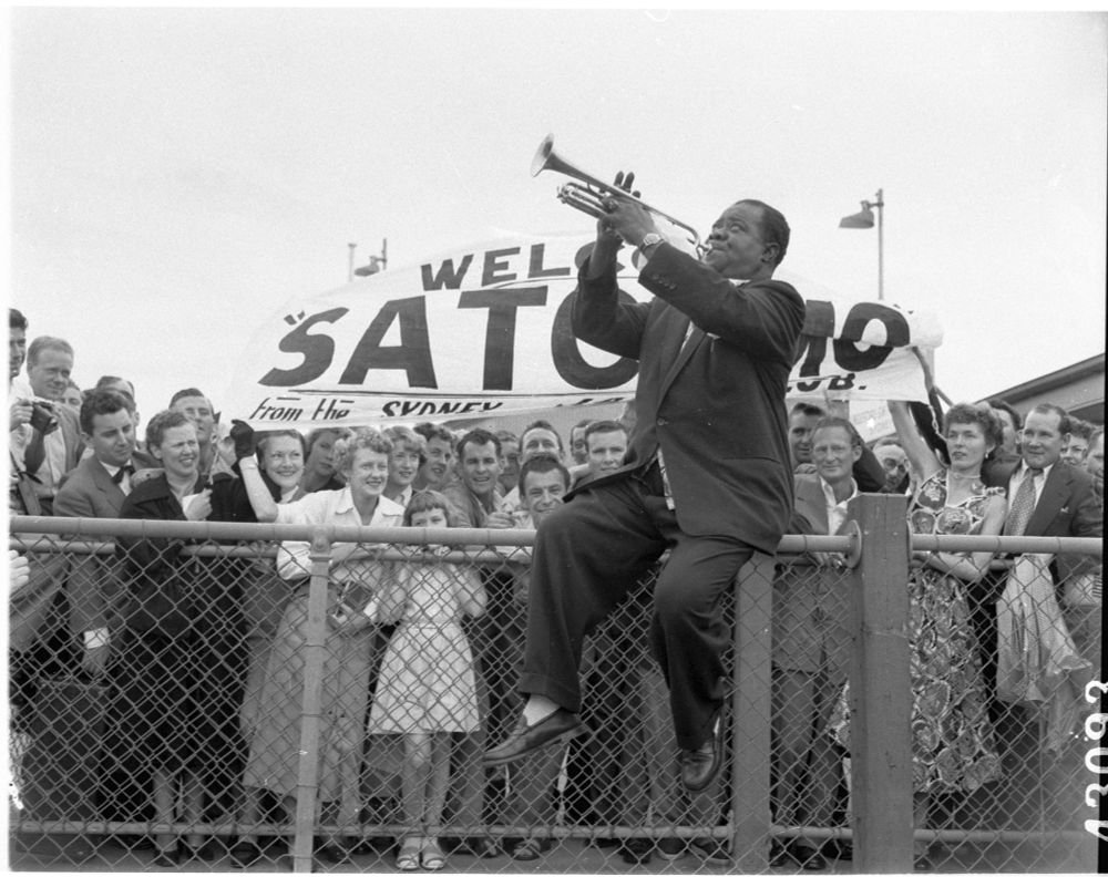 A photo of Louis Armstrong arriving in Sidney, Australia, in 1954. He is sitting on top of a fence, dressed in a suit and playing his trumpet. There is a crowd of people gathered behind him, with some holding a large banner that says "Welcome Satchmo" 