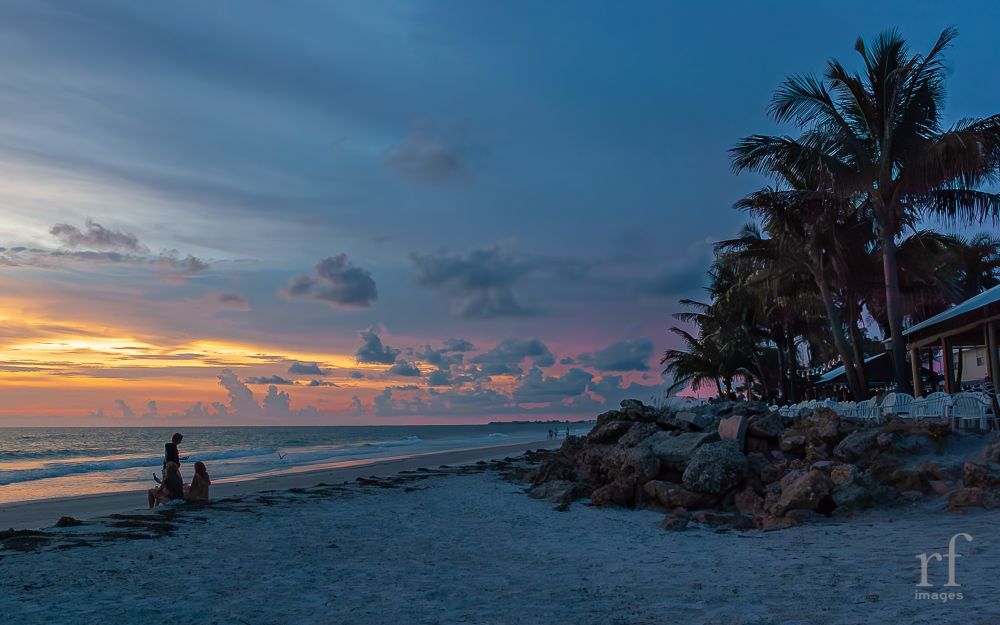 A couple enjoys the beach at sunset. Magenta, orange and yellow streak the sky but can’t overcome blow hour shadows on the people, rocks, and palm trees. 