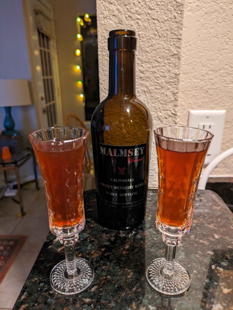 A bottle of Malmsey on a dark granite countertop, with two full dessert wine glasses to the left and right. String lights softly glowing in the background.