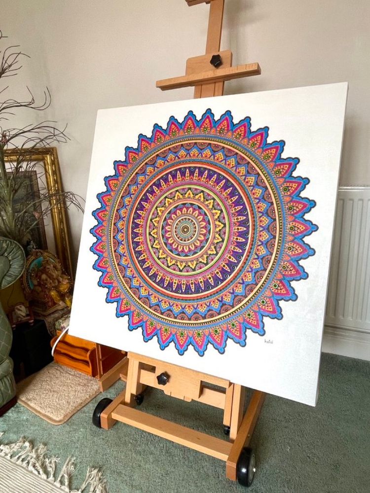 A detailed painted mandala in brights colours on white background sits on a h frame easel in a room with a green carpet.