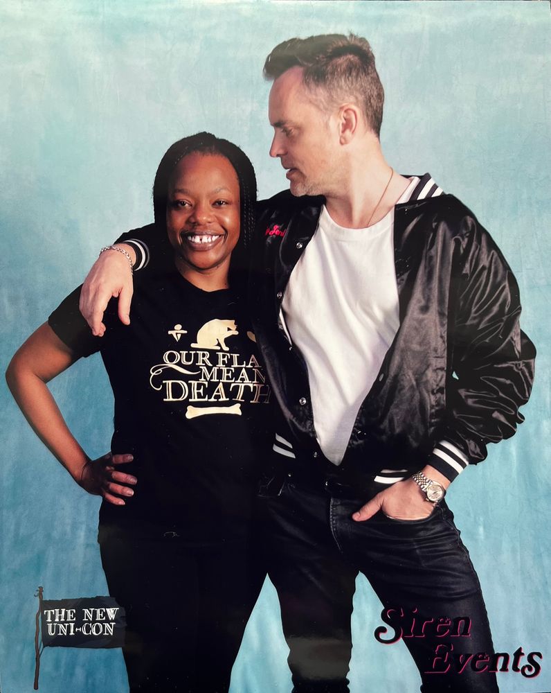 One person looking forward wearing an Our Flag Means Death t-shirt and black pants. Their hand is on their hip. The second person looking at the first person wearing a white t-shirt, a black jacket and black pants. They have one hand in their pocket and their second hand over the first person’s shoulder. Writing on the bottom of the picture says The New Uni-Con on the left hand side and Siren Events on the right hand side.