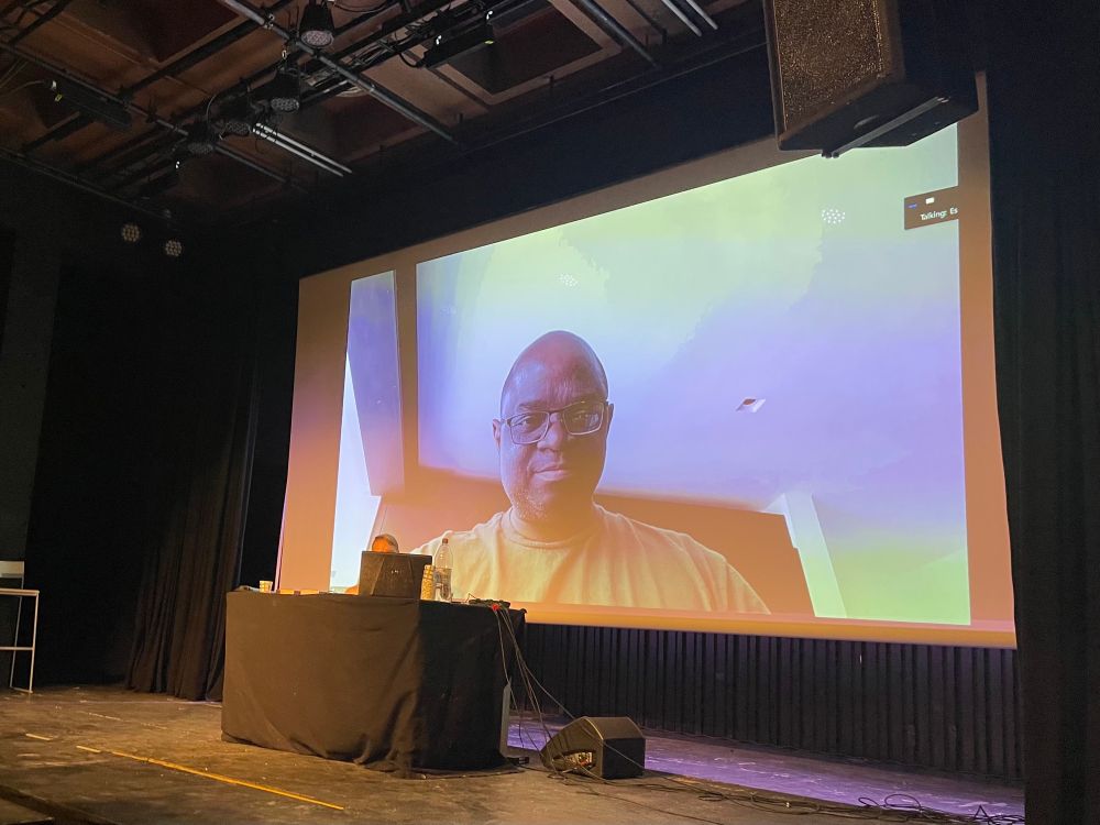 Tade Thompson interviewed via zoom on a Big screen, with interviewer Esko M. Suoranta being small in front behind a laptop.