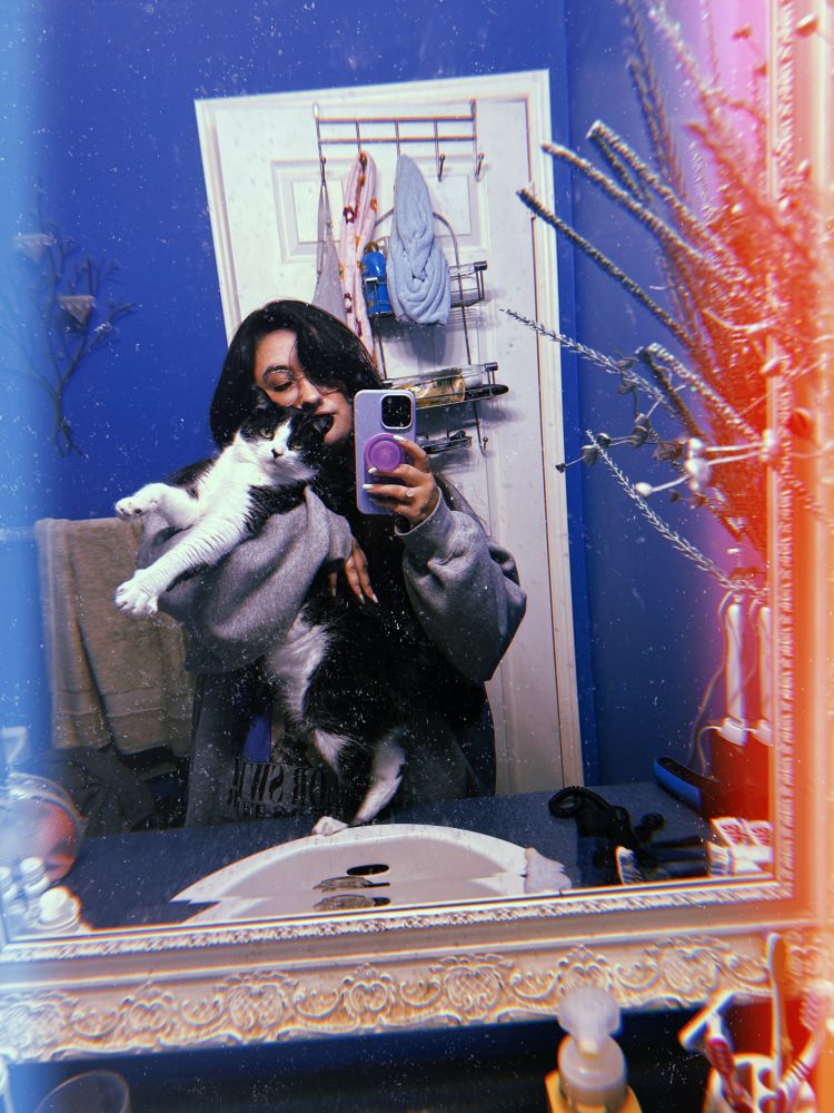 Shelby from summer 2024 in my old bathroom. I’m taking a mirrror selfie with my male cat Moe, he is a black and white cat. I am holding him with my arm and his face is turned towards the mirror 