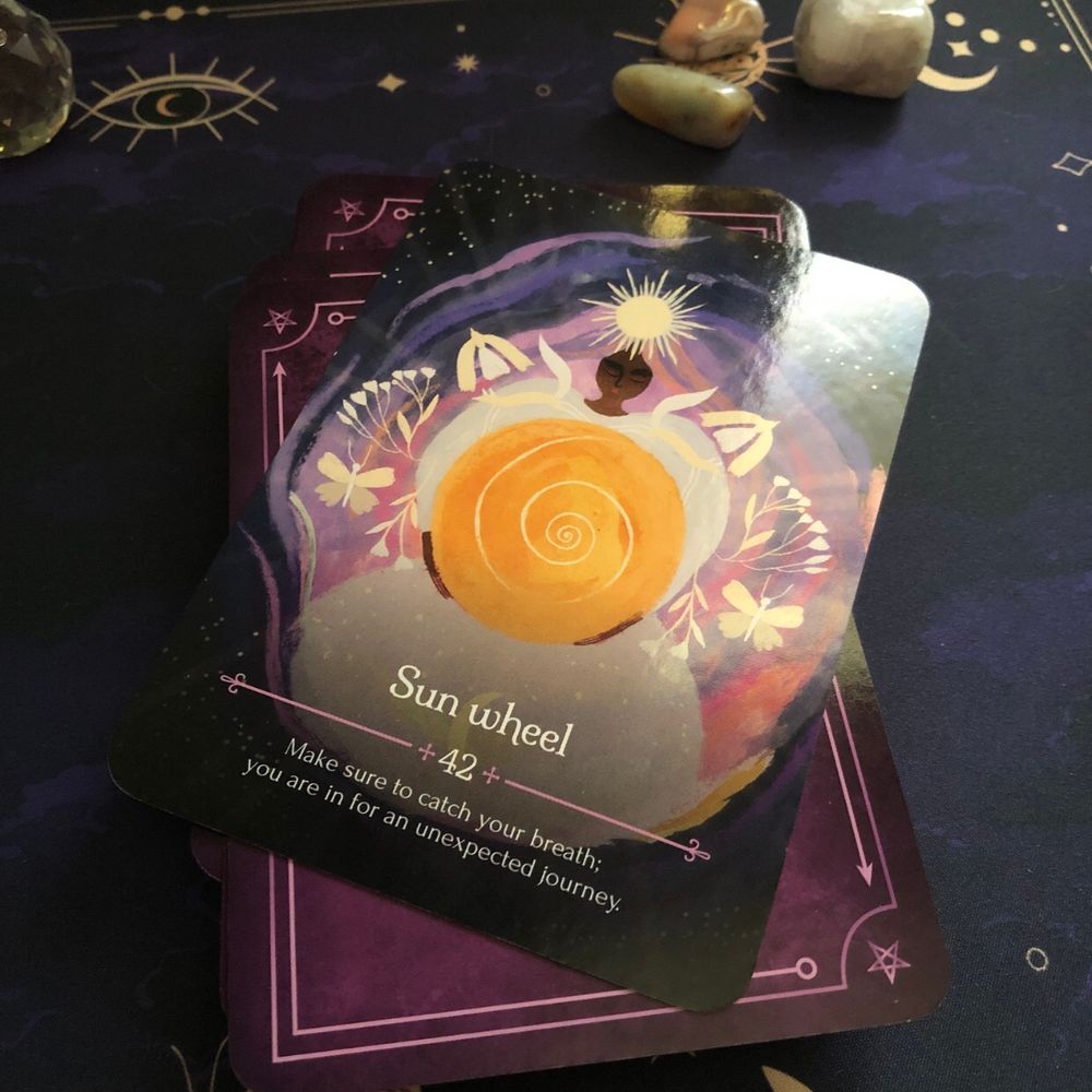 The Sun Wheel card from the Seasons of the Witch Litha Oracle deck. The card shows a beautiful pale clothed figure holding a spinning yellow circle. The card’s background is purple starry sky, moths and flowers flutter next to the figure. The card says: Make sure to catch your breath; you are in for an unexpected journey. 