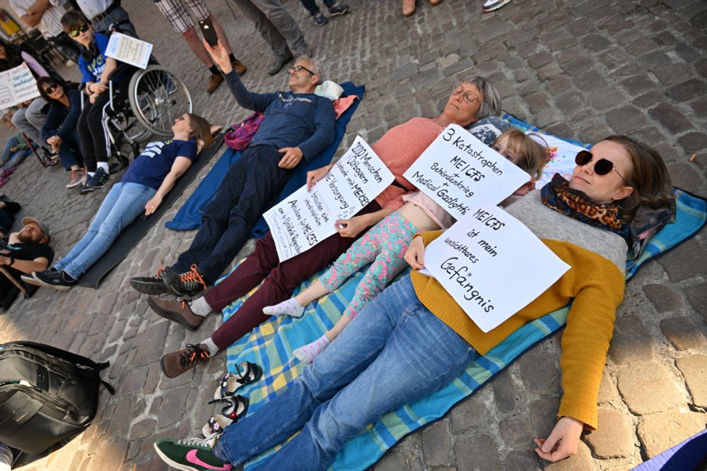 Fünf Personen liegen auf Matten am Boden, eins davon ist ein Kind. Ein weiteres Kind sitzt daneben im Rollstuhl. Drei der liegenden Personen haben ein Schild auf ihren Körpern liegen. Auf dem ersten steht "ME/CFS ist mein unsichtbares Gefängnis". Auf dem des Kindes steht "Drei Katastrophen - ME/CFS plus Behördenkrieg plus Medical Gaslighting". Auf dem dritten steht "20.000 Menschen in Ostbayern schwerkrank mit ME/CFS ohne Versorgung." Daneben sind Tränen eingezeichnet. Darunter steht "Wir brauchen eine interdisziplinäre Ambulanz für ME/CFS an der Uniklinik Regensburg - Therapie, Heilversuche, Studien"