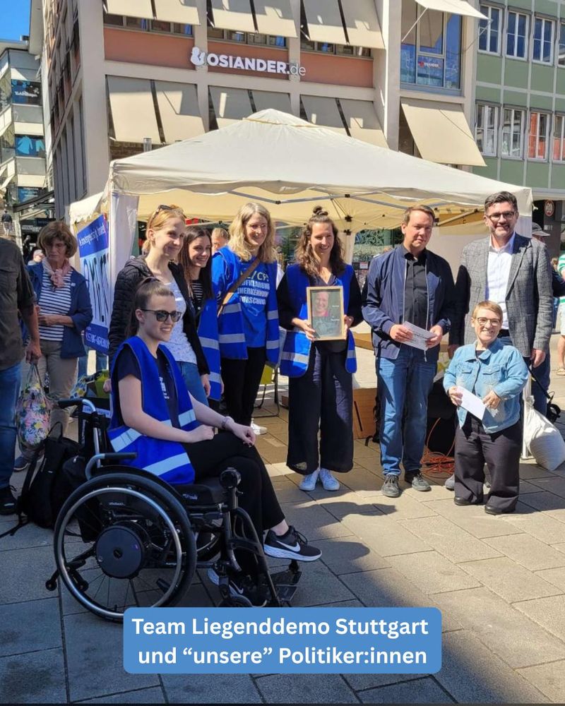 Vor einem Infostand stehen mehrere Personen - Ordner:innen mit blauen Warnwesten und Politiker:innen. Eine Person sitzt in einem Rollstuhl. Ein Text beschreibt das Bild mit den Worten "Team Liegenddemo Stuttgart und "unsere" Politiker:innen"