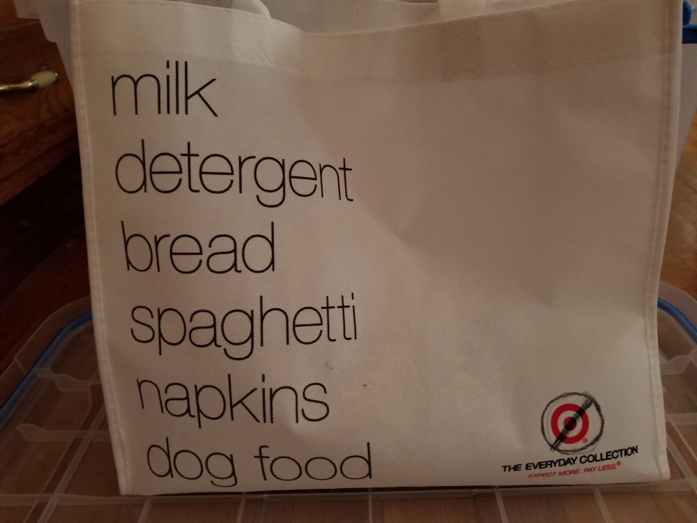 A white reusable shopping bag. In large black letters, it lists, "milk, detergent, bread, spaghetti, napkins, dog food". In the lower right corner is the Target logo, with the words "The Everyday Collection" and the store slogan " Expect more, pay less" underneath it. A black circle with a slash has been drawn over the logo.
