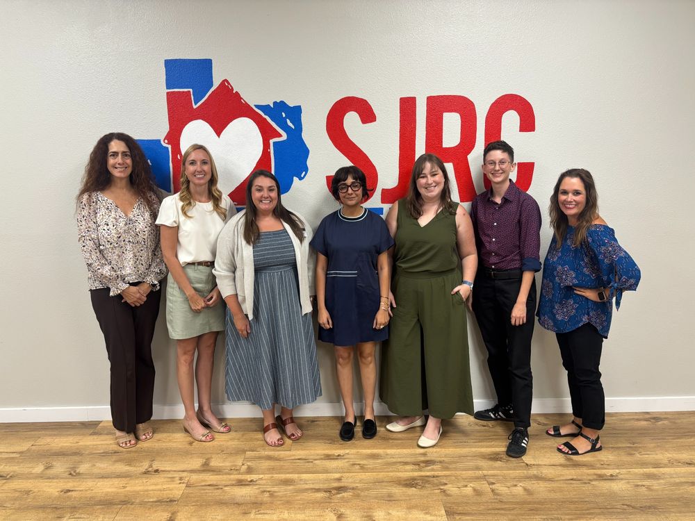 Seven people smiling for the camera in front of a wall with the SJRC logo.