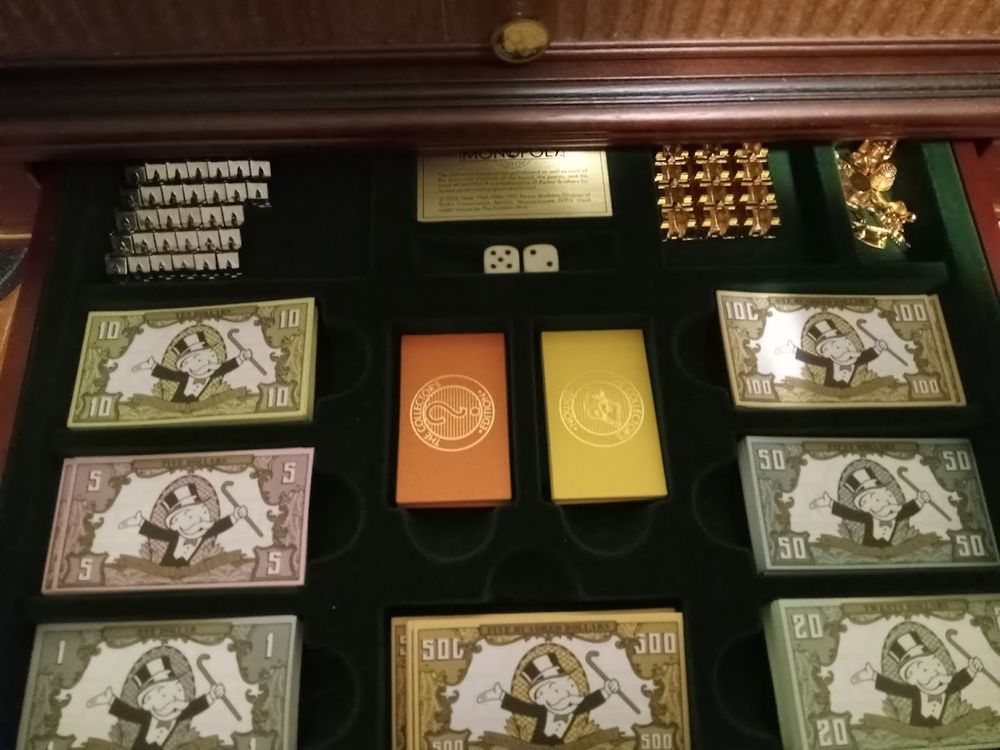 A drawer from a Monopoly table displaying two dice, chance and community chest cards, Monopoly money, silver houses, gold hotels, and gold playing tokens.