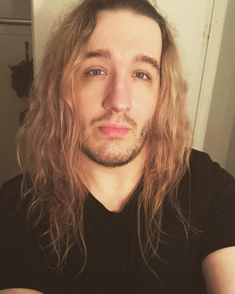bathroom selfie of me from late 2022, with long, wavy blonde hair and a short beard, wearing a black v neck t-shirt
