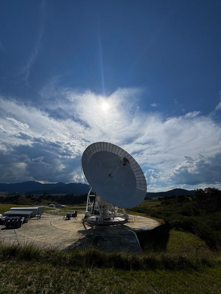 Sun shining through clouds in a blue sky above a mountainous region with a deep space antenna in the foreground.