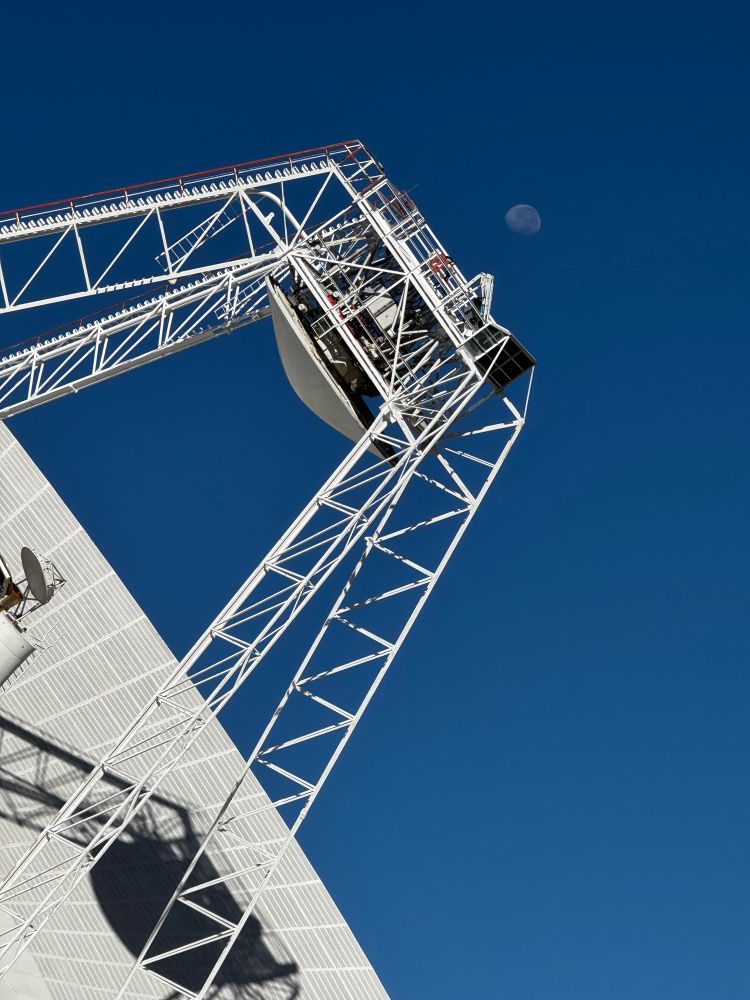 Daytime image of the Moon above the metal structure of a deep space communications antenna dish.