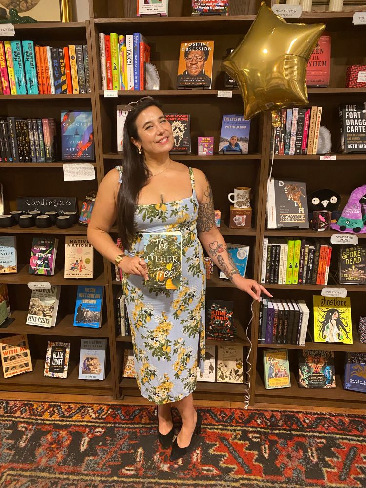 alison in a blue dress with lemon print holding up their new award-winning book, THE OTHER TREE, whose cover matches the dress 