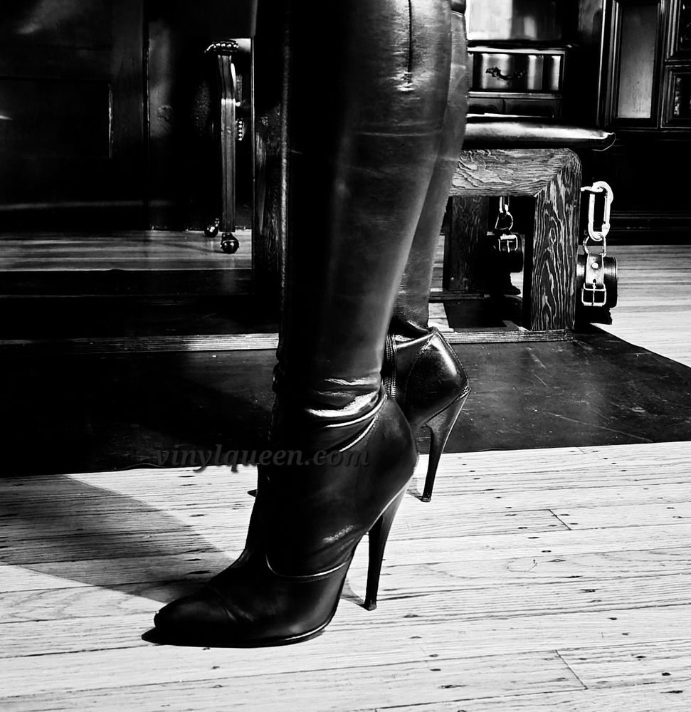 Mistress Vinyl Queen's leather boots in black and white on a wooden floor from the knees down.