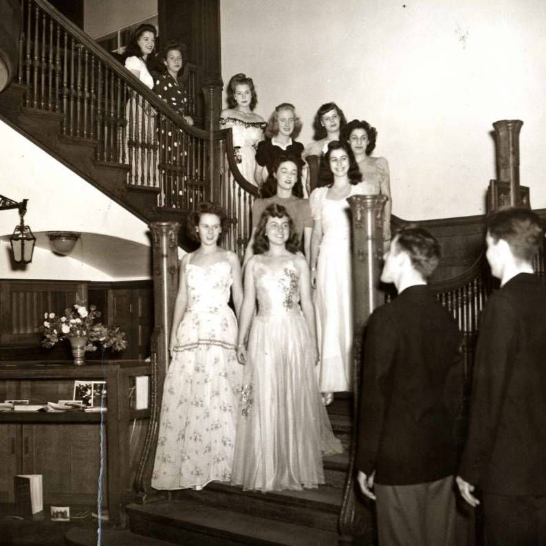 college students get ready for a formal social. the women stand on a staircase, wearing floor-length dresses. the men stand at the foot of the steps, wearing blazers.