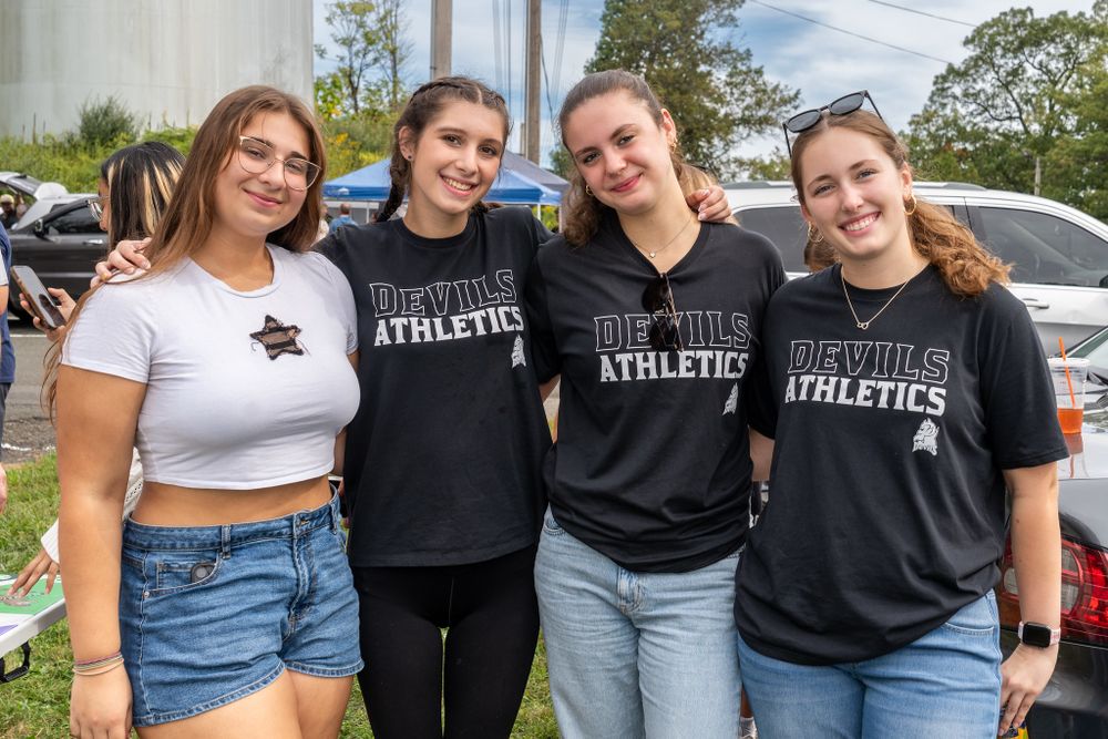college students stand in a parking lot and smile at the camera. three of the four students wear FDU Devils Athletics shirts.