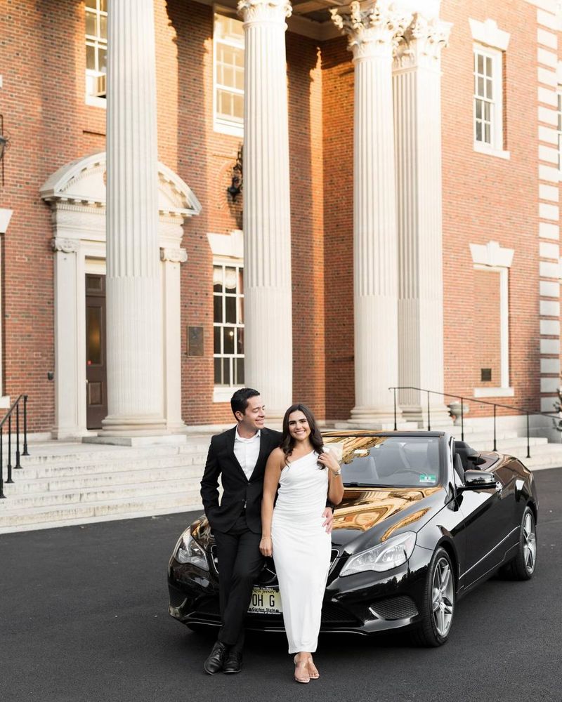 a couple holds hands and smiles at the camera. they lean on a car outside of an estate. the woman wears a white dress and the man wears a black suit.