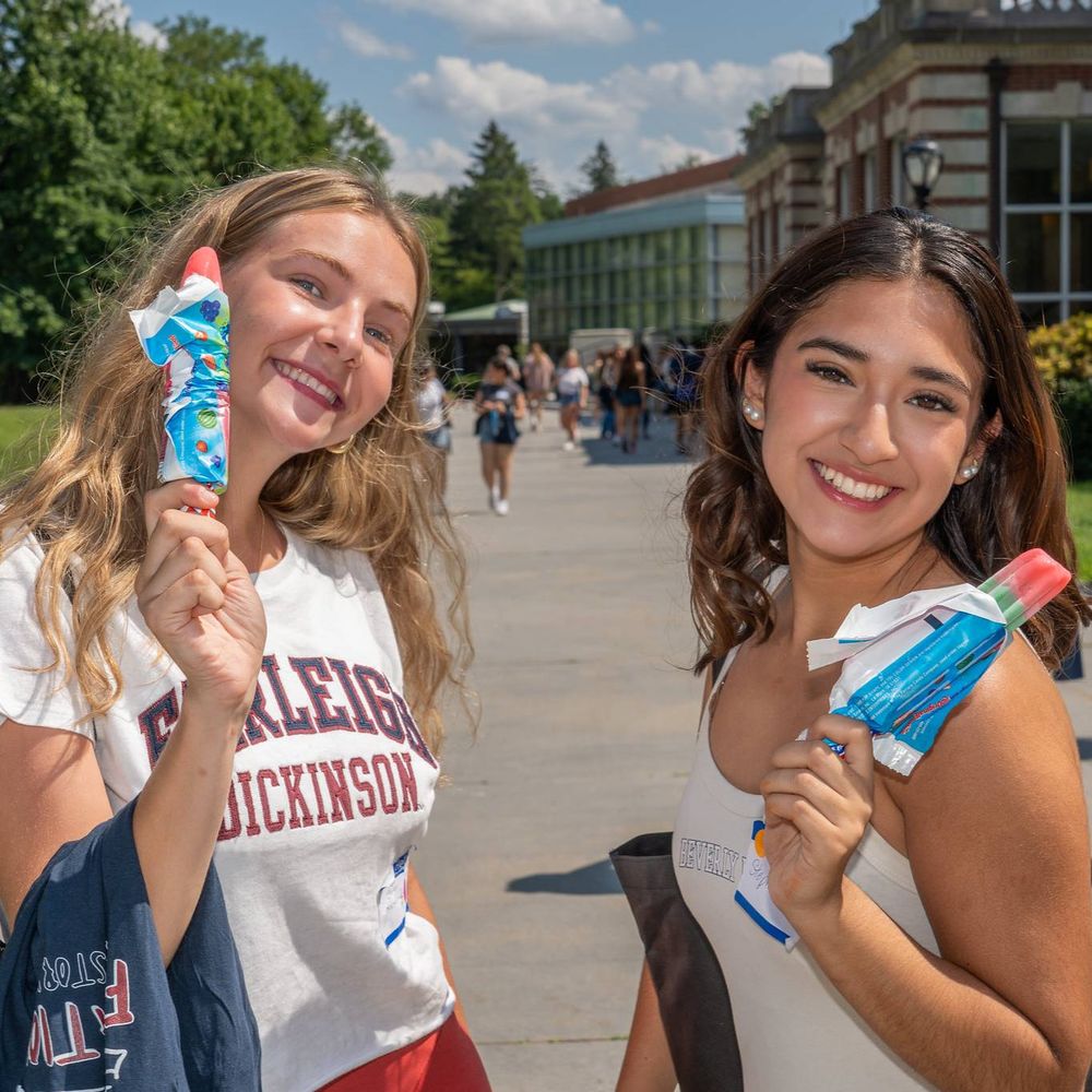 two FDU students hold ice cream and smile at the camera. one wears a tank top and the other wears an FDU shirt. they stand outside as students walk in the background.