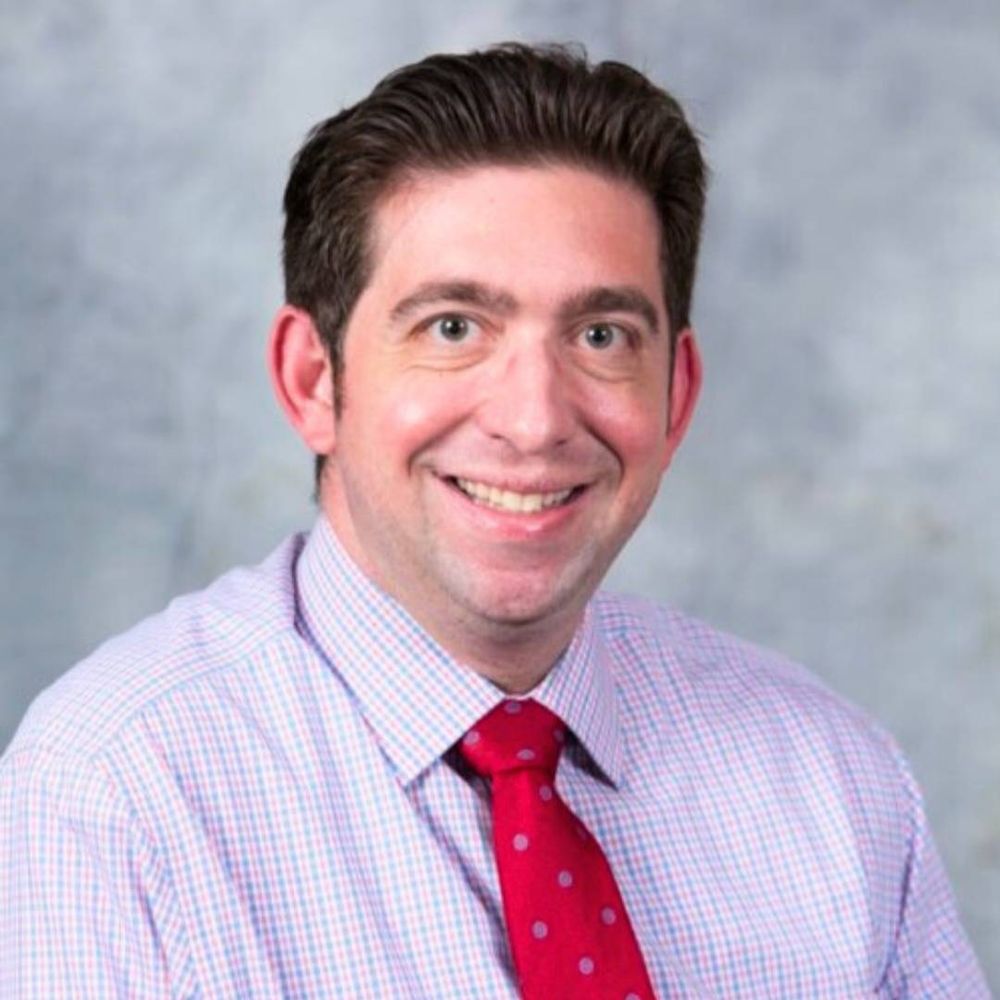 portrait of a man against a gray backdrop. he wears a red tie.