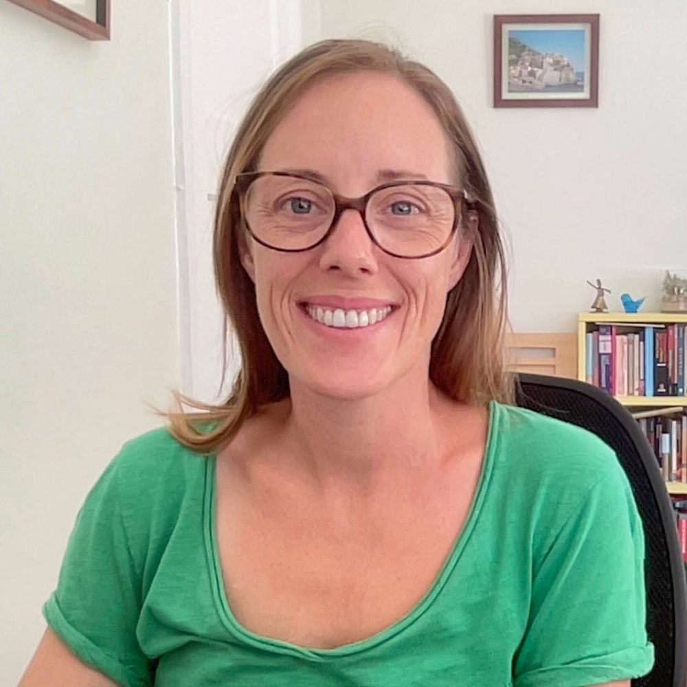 a headshot of a woman. she wears a green shirt and there is a bookshelf and framed photo behind her.