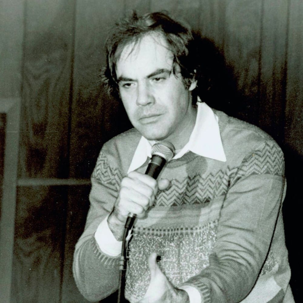 Robert Klein holds a microphone and speaks to an audience of college students that are out of frame of the photo. he wears a sweater.