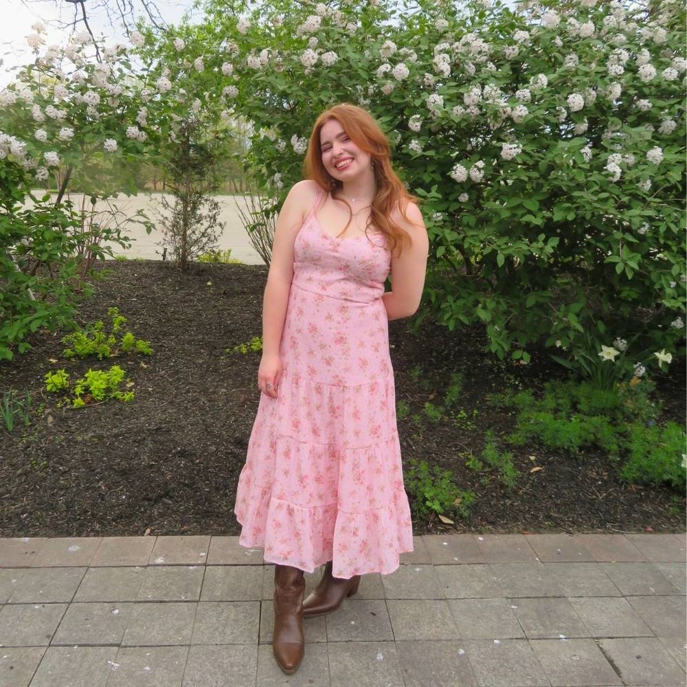 a university student stands outside and smiles at the camera. she wears a pink dress and is surrounded by flowers.
