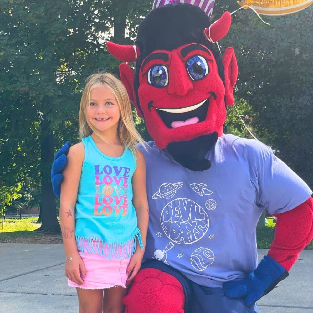 the Ian the Devil mascot squats down next to a child. they stand outside and smile at the camera.