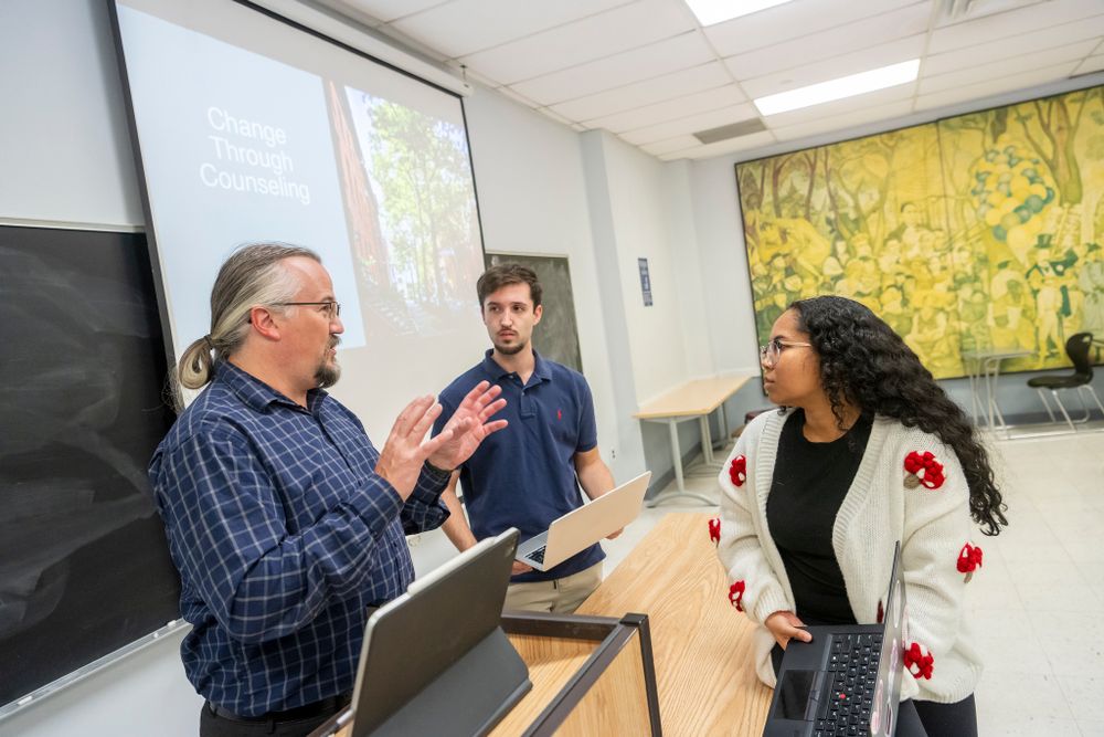 students chat with a professor. they stand around a desk with their laptops open. the presentation behind them reads "change through counseling."