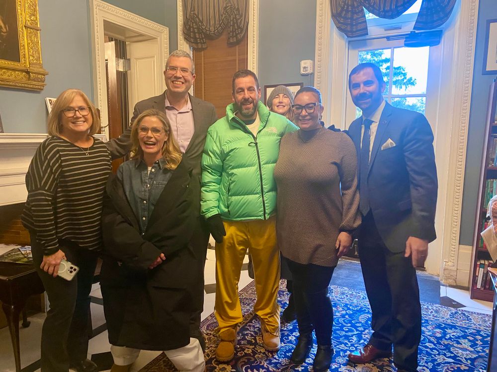 Adam Sandler, Julie Bowen and FDU President Michael Avaltroni stand with a group. They smile at the camera.