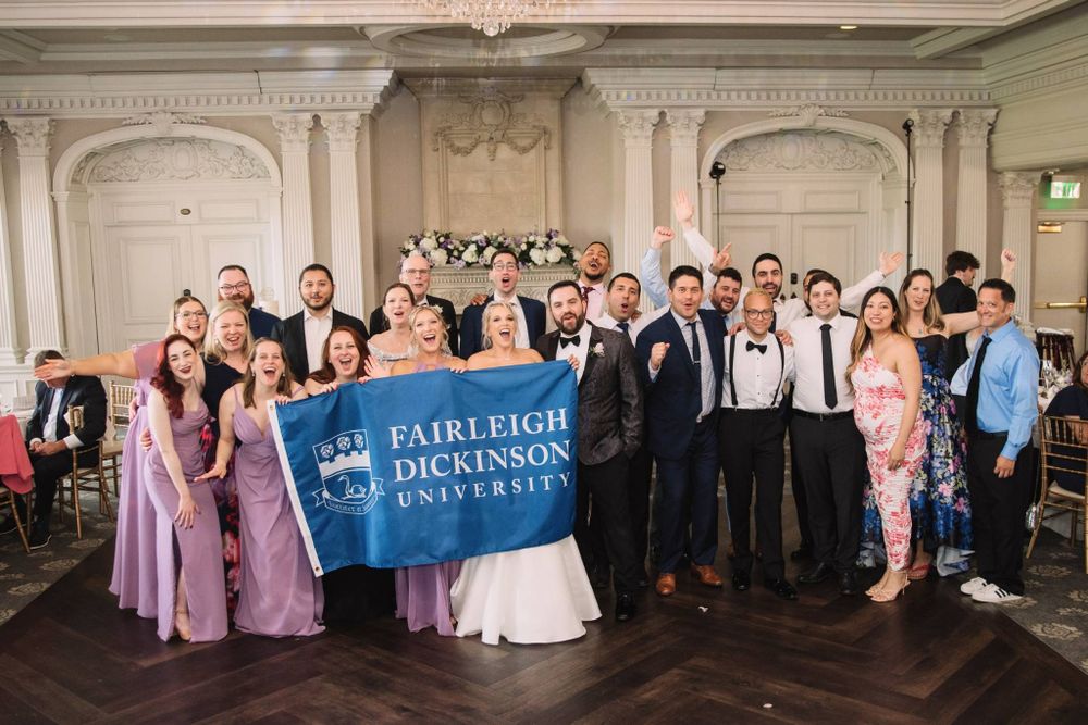 a bride and groom stand in the middle of a group. they hold up a banner for FDU. the group around them cheers.