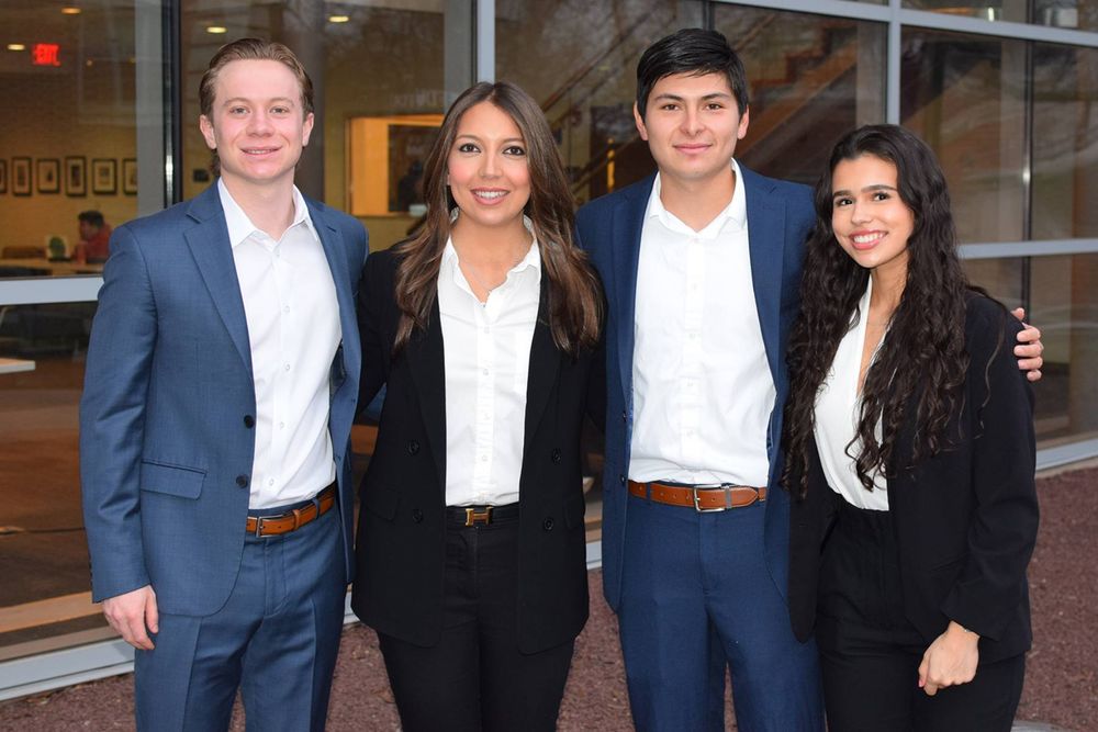 Four students stand in a line, posing for a photo. Two are male, two are female. they wear business attire.
