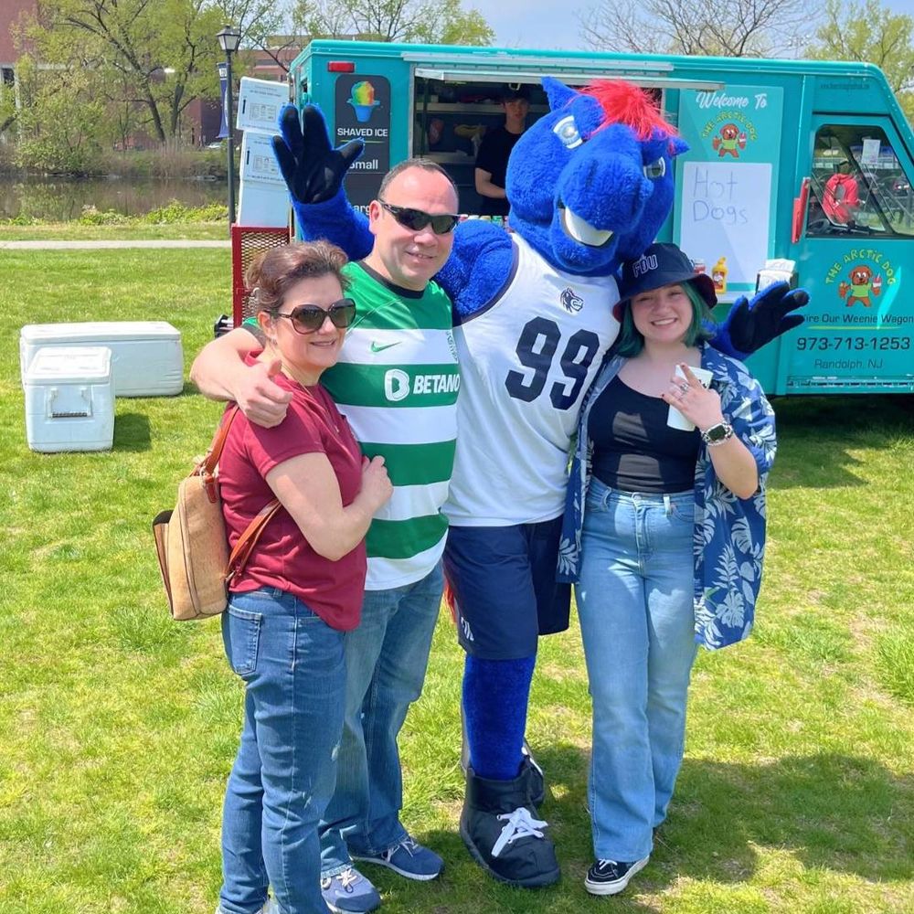 the Knightro mascot stands with a group of people. one wears an FDU Knights bucket hat. there is a food truck behind them.