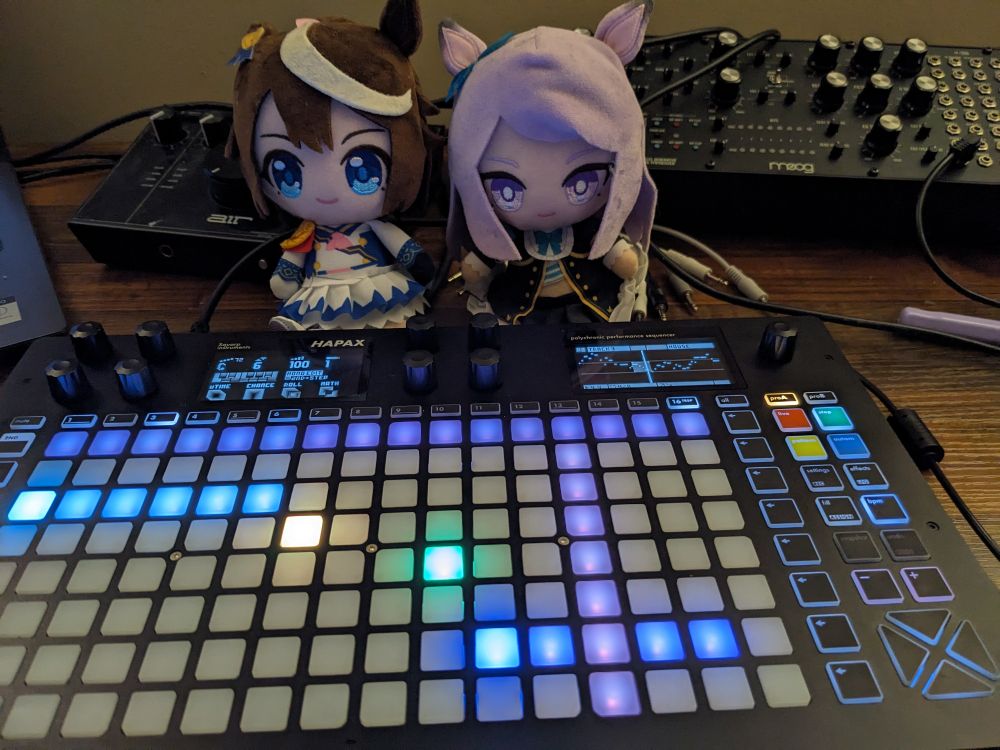 picture of music studio desk with laptop, audio interface (cheapo quality), moog labyrinth (still working this one out). two plushies of umamusume characters (Tokai Teio and Mejiro McQueen) sit behind a squarp instruments hapax sequencer , the focal point of the image. it has glowy lights and a lot of pads and knobs