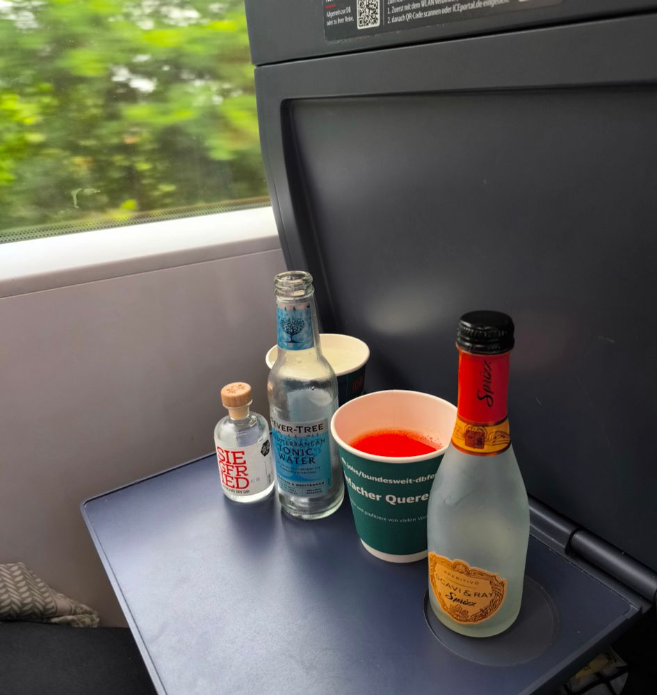 Blauer Klapptisch im ICE, darauf (von vorne nach hinten) ein leerer Piccolo Scavi&Ray Sprizz, ein orange gefüllter Pappbecher, eine leere Flasche Tonic Water, Miniatur-Flasche Gin, ein transparent gefüllter Pappbecher. Im Hintergrund ein Teil des Zugfensters, draußen verschwommenes Grün. 