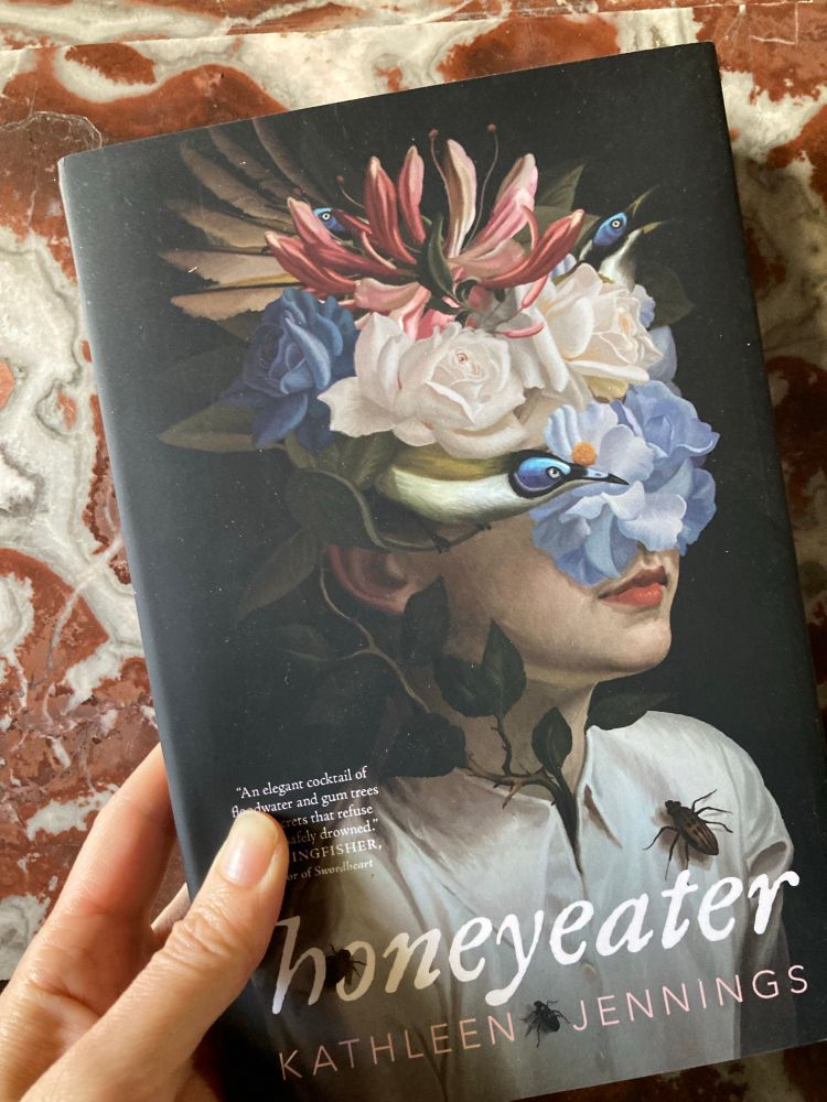 My hand holding a copy of Honeyeater by Kathleen Jennings in front of red marble. The cover illustration shows a young woman in a white blouse with most of her face obscured by a headpiece of tropical flowers, insects on her blouse. Like a Golden Age Dutch still life but with a person in it