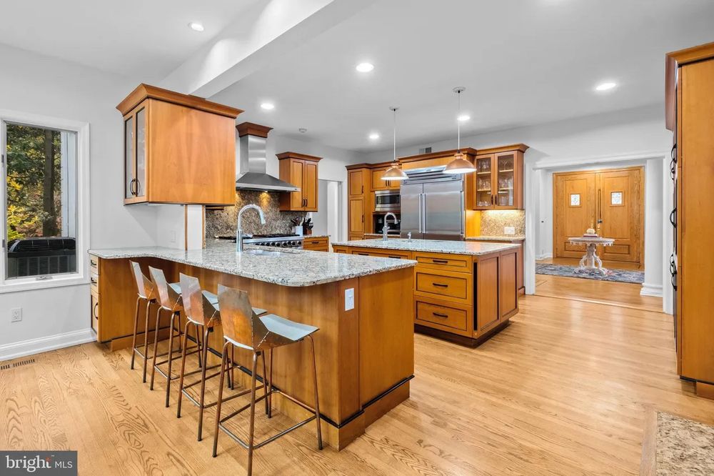Kitchen with hardwood flooring, granite counter tops, two islands; each with a sink. A huge stove with a hood vent. Seemingly a large oven and an in wall oven. A huge fridge and freezer. All the cabinets are wood with metal finishings. 