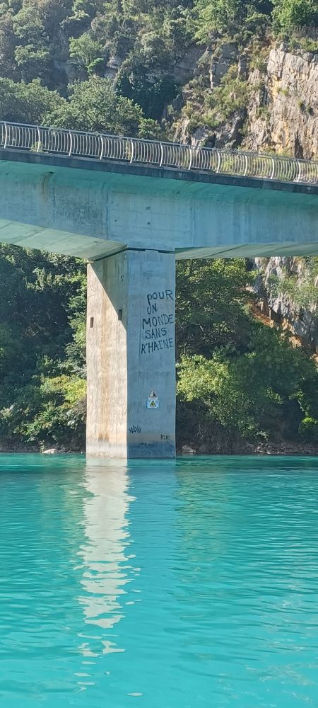 Pile d'un pont avec un graffiti qui indique "pour un monde sans R'Haine".
Eau très bleue sous le pont