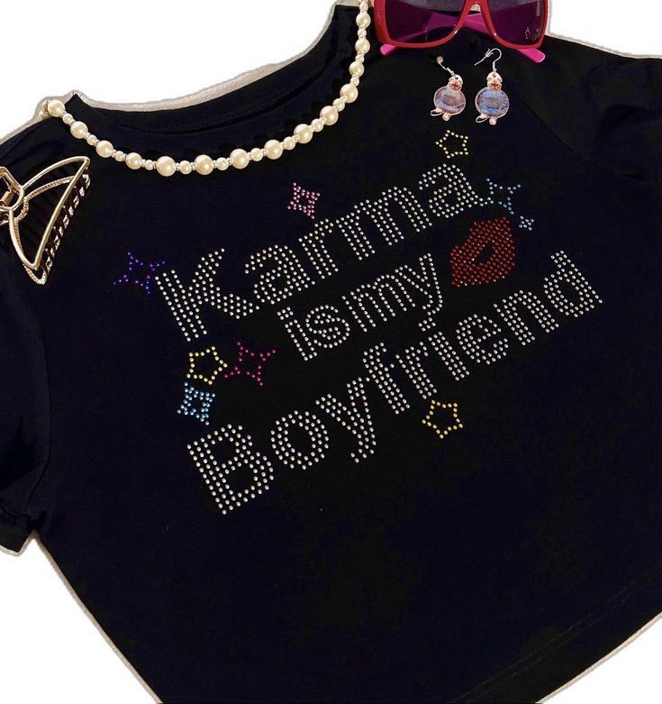 A black, short sleeved crop top. “Karma is my Boyfriend” is written in silver rhinestones. There are also rhinestones making a red lip and stars in the following colors: yellow, pink, purple, and red. There are also the following accessories placed by the top: a gold hair clip, a pearl necklace, pink sunglasses, and earrings that I can’t quite make out. I can tell that they are pink, dangly, and have a spherical shape with attatchments, but I have no idea what they are supposed to resemble.