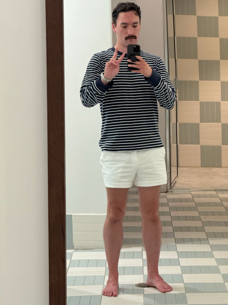 A barefoot man wearing a pair of white shorts and a blue and white striped long sleeved shirt poses in front of a gym locker room mirror. 
