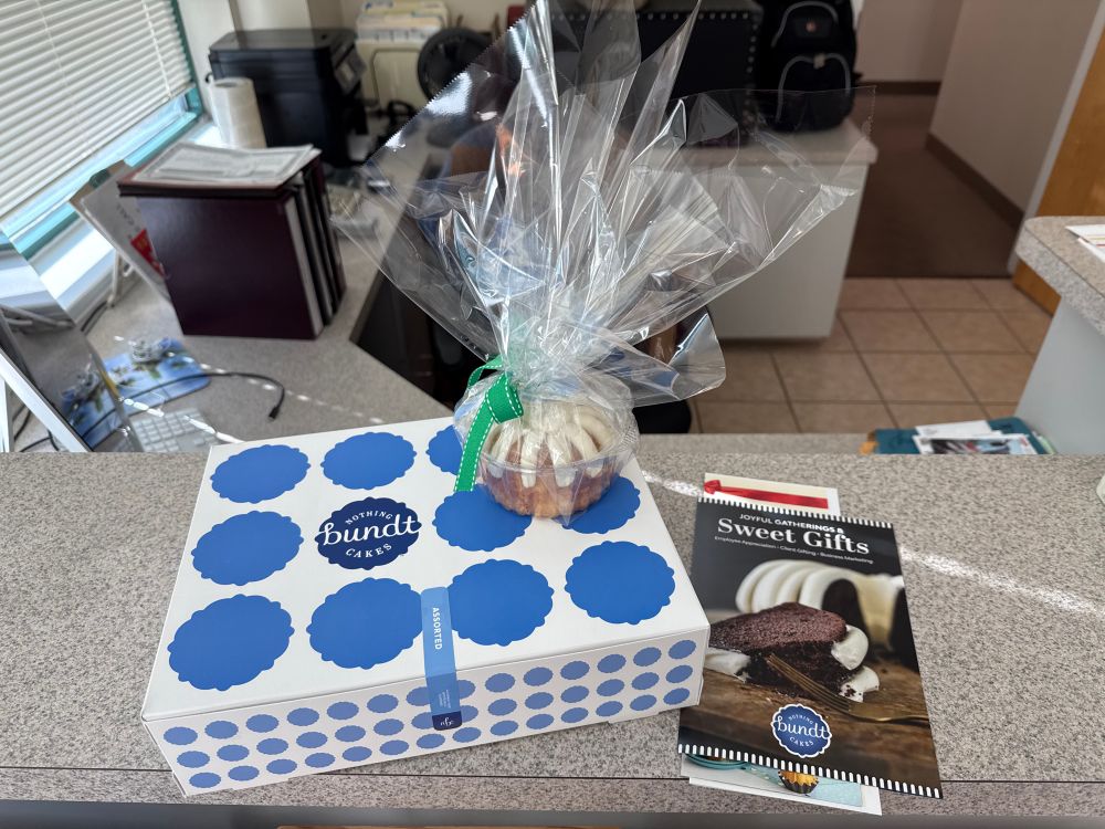 A white cardboard box with blue dots sits in a countertop, with a vanilla Bundt cake sits atop wrapped in clear plastic. 