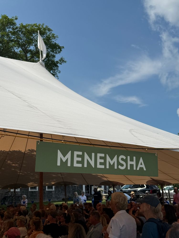 A green sign with white lettering that says MENEMSHA is affixed to a canvas tent. A crowd of panel attendees sits and stands under it. 