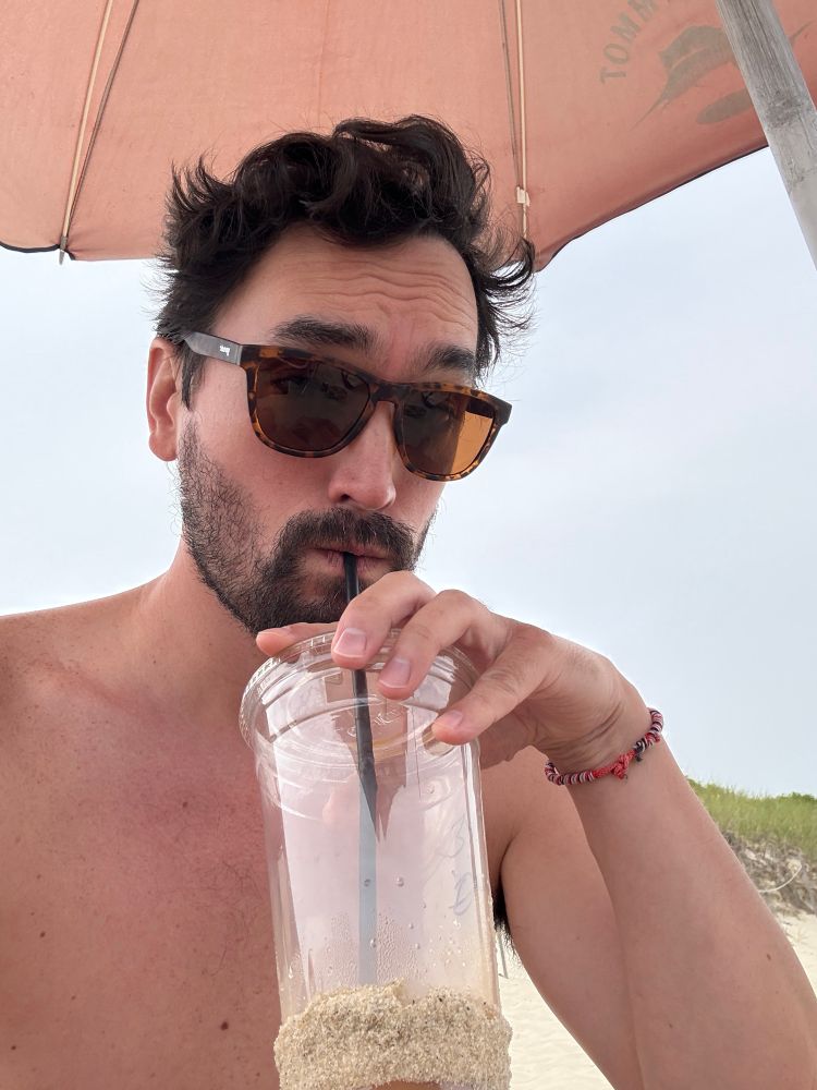 A shirtless man sits underneath an orange beach umbrella and sips out of a plastic cup whose bottom is covered in sand. 