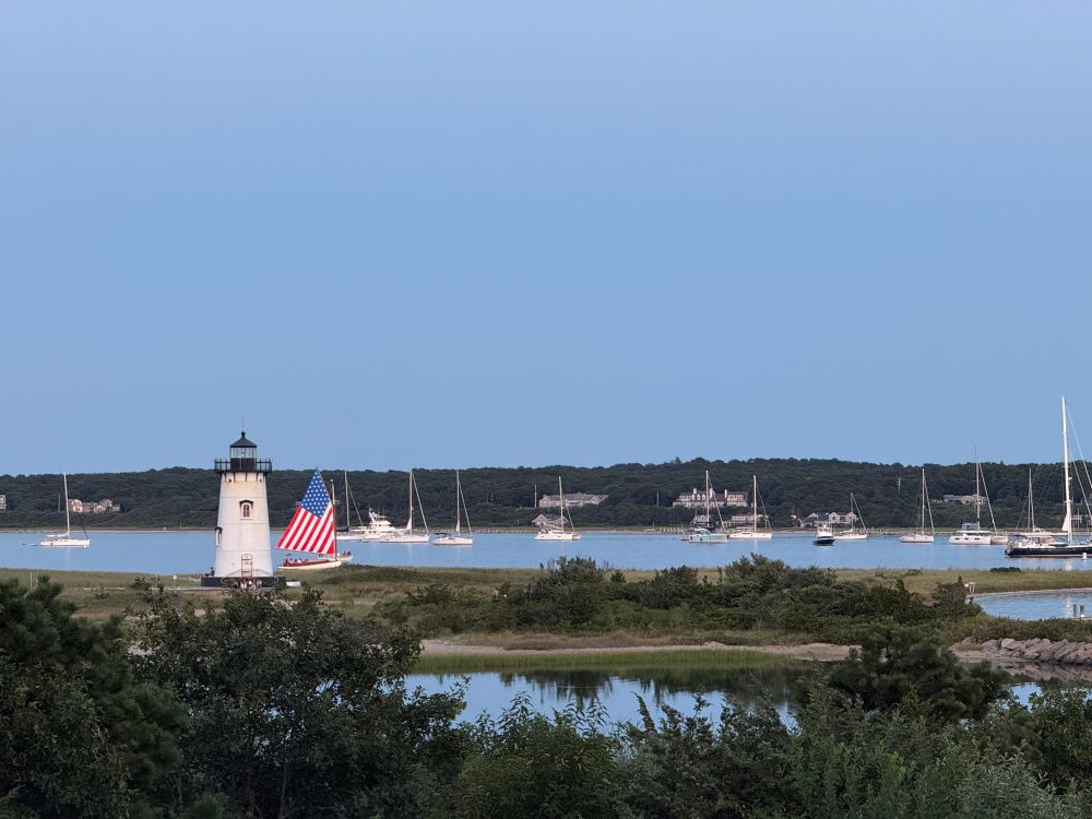A white lighthouse guards an inlet filled with anchored sailboats. A boat with an American flag as its sails glides past. 