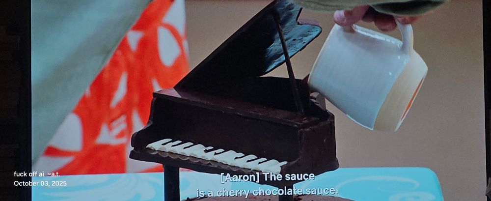 A small piano, perfect imitation, made entirely of chocolate. It probably about two hand spans high and wide. The top is opened and someone is pouring chocolate sauce into it for tasting. 