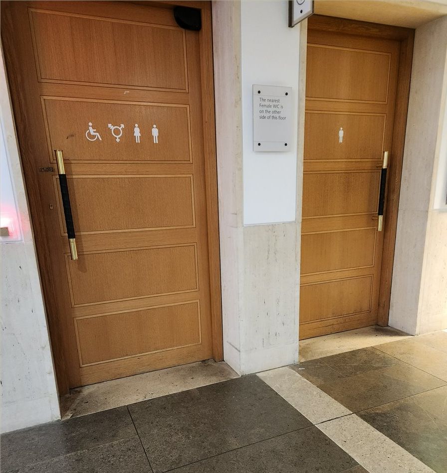 Photo of two wooden doors with long tall handles for pushing open. There is a hard concrete clean floor in front of them. Door on left has four symbols from left to right: handicap wheelchair, three pronged gender circle, standard bathroom figure with dress for female, standard bathroom figure with pants for male. In contrast, door on right has only one symbol: figure with pants for male. 
