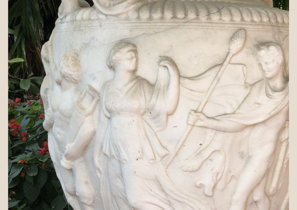 Close up photo of Greek-style figures sculpted onto the exterior of a large vase sitting in a garden. 