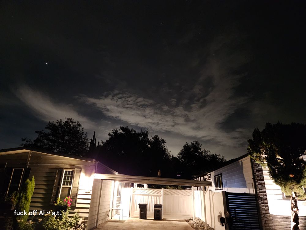 Night shot, cloudy sky with clouds that look like a huge pale bird flying over some dark trees. In front bottom of photo is a lit up driveway for a little one level house with plants by the front windows. It is a dark night and no people are out. 