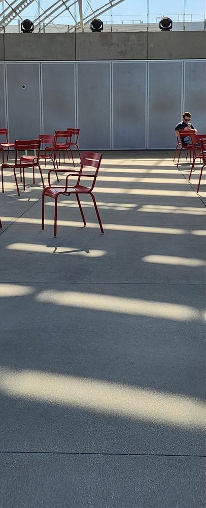Very tall narrow photo, similar in shape to a long bookmark. A majority of the photo is a concrete patio covered in slashes of light from a slotted roof above that we cannot see. A number of red metal chairs are scattered about a large space. There is a concrete wall at the far back top of the image. There is only one person in the image: at far upper right, sitting in the furthest back of the metal chairs. Short hair, wearing sunglasses, blue shirt is about all you can make out - and the fact that their head is bent down as they look at an object they are using both hands to tap on (which is a smart phone, per the photographer knowing it is, but otherwise you'd be guessing that's what they were busy using all alone in this empty space).