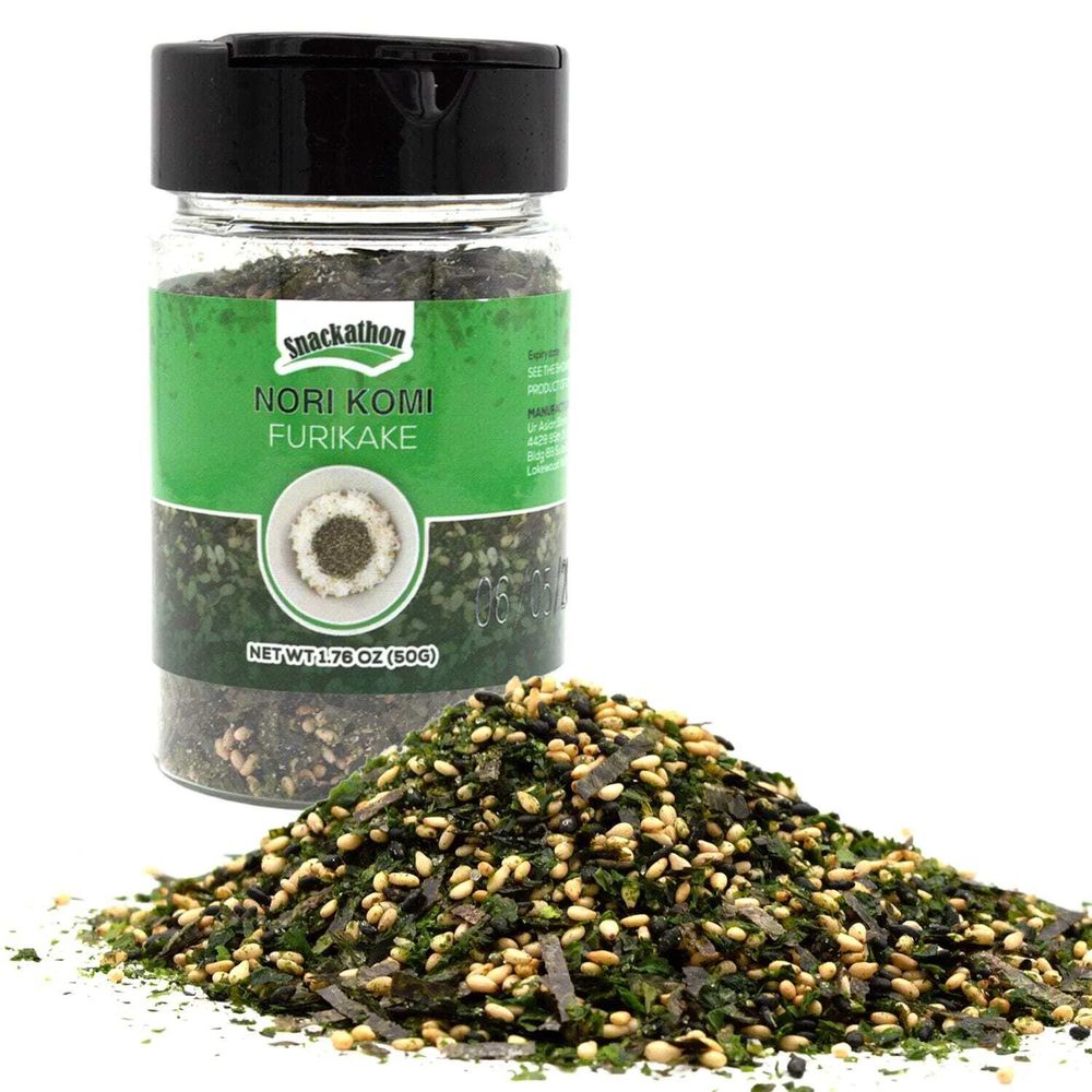 Photo of a screw top bottle with a label on the side that reads: "Nori Komi Furikake". In front of the bottle is a pile of furikake, which has a combination of tiny flakes of green and pale roasted bits seaweed.